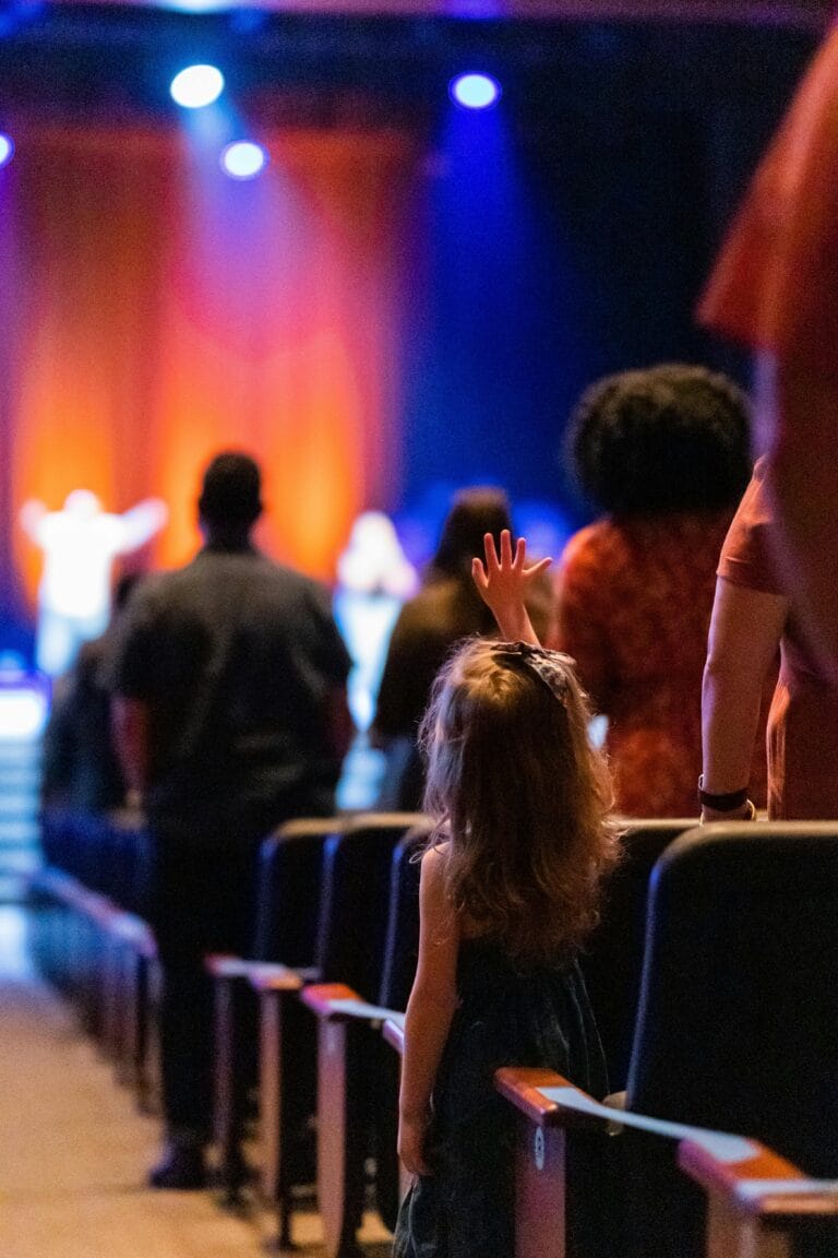 pexels photo 7626321 7626321 reserva teatro para colegios - A young girl stands amidst a vibrant audience during a live concert indoors.